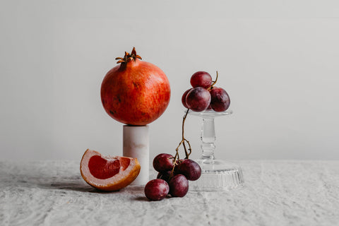 Grapes on an upturned glass next to a slice of pomegranate and a whole pomegranate on a marble stand, against a white background