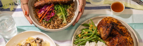 Hands holding a Joel's place rice bowl meal next to two plates of Joel's Place chicken and pasta dishes, a glass of water, and dip, on top of a striped tablecloth