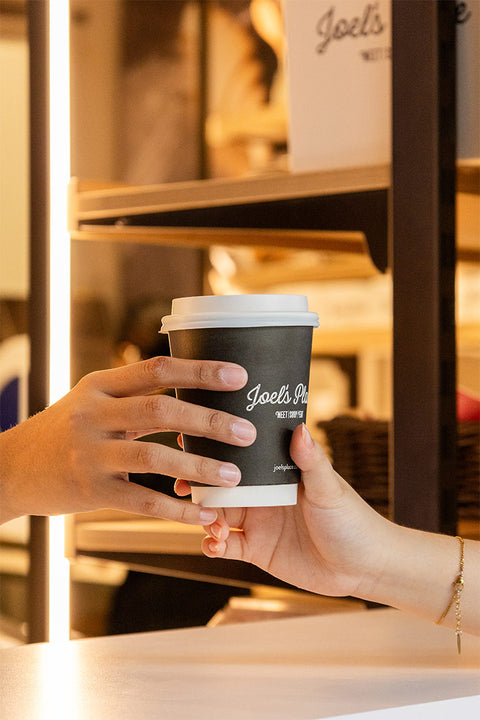 A close-up shot of two hands, one reaching out to receive a coffee cup with 'Joel's Place' logo from another hand.