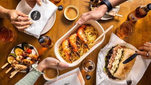 A flatlay shot of a group of people eating diverse takeout meals spread on a wooden table, featuring pasta, sandwiches, skewers, and beverages from Joel’s Place Click and Collect.