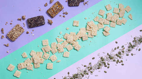 A variety of snacks are lined up in slants against purple, light green, and pink background. It features crisps, nuts, and granola bars.