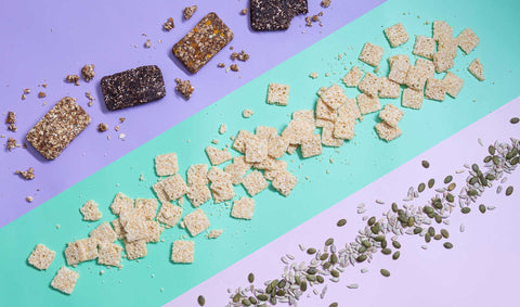 A variety of snacks lined up in slants against purple, light green, and pink background. It features crisps, nuts, and granola bars. 