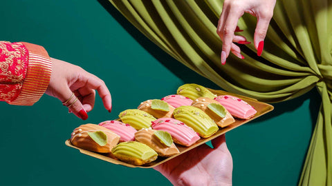 Two hands aiming to pick up Joel's Place madeleines perfectly lined up in a gold tray with flavors like Earl Grey, Pistachio, and Raspberry. 