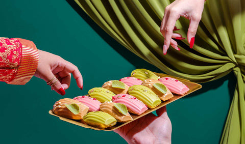 Two hands aiming to pick up Joel's Place madeleines perfectly lined up in a gold tray with flavors like Earl Grey, Pistachio, and Raspberry. 