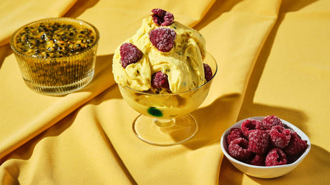 A cup of Passion Fruit & Raspberry Gelato  by Chef Miko placed on top of a yellow cloth with small bowls of passionfruit and raspberries on the side.