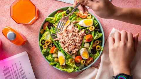 A hand holding a fork over a Joel's Place salad, next to a glass of orange juice and a power shot bottle, on a pink tabletop