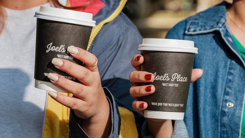 Two people standing side by side outdoors, each holding a black takeaway coffee cup of Joel’s Place. One person wears a yellow and blue jacket with nude-colored nails, while the other wears a denim jacket with red nail polish.
