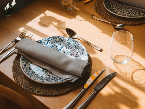 A formal dining setting for two featuring blue floral plates and circular woven placemats, on a wooden tabletop
