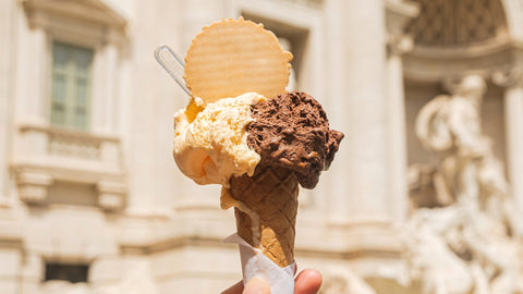 An ice cream cone with two flavored scoops of gelato and a thin waffle on top with plastic spoon peeking on the side. It is against the backdrop of the Trevi Fountain. 