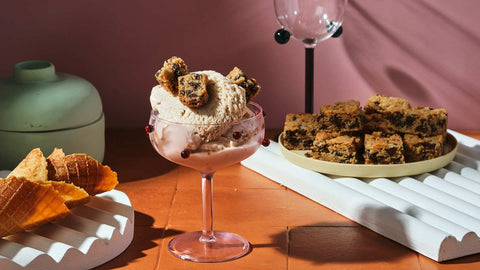 A cup of New York Cheesecake and Cookies Gelato by Chef Miko against a pink background with several cones on the left and square bite-sized cookies on the right. 