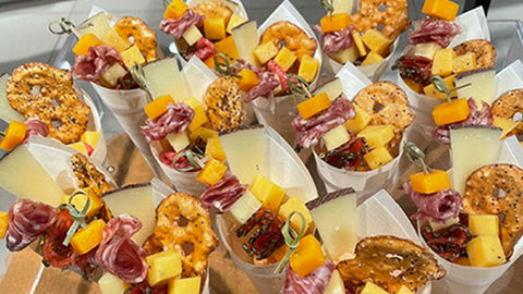 Several white paper cones filled with assorted cheeses, cold cuts, and pretzels