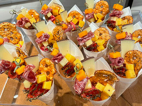 Several white paper cones filled with assorted cheeses, cold cuts, and pretzels