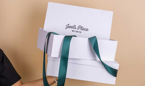 A person on the side holding a stack of white Joel's Place boxes with green ribbon. 