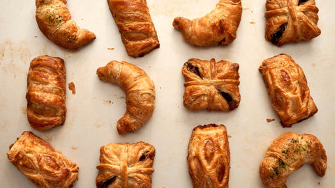This image displays a flatlay of freshly baked, laminated dough pastries, characterized by their golden, flaky, and buttery layers, that include cronadas, empanadas, and filled pastries,  available at Joel’s Place. 