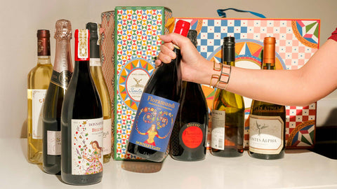 A selection of wine bottles arranged on a countertop, with a colorful wine gift box in the background. A hand is holding a bottle of Floramundi wine by Donnafugata