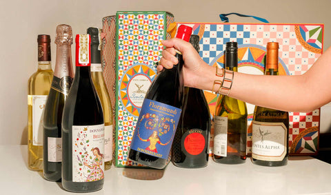A selection of wine bottles arranged on a countertop, with a colorful wine gift box in the background. A hand is holding a bottle of Floramundi wine by Donnafugata.
