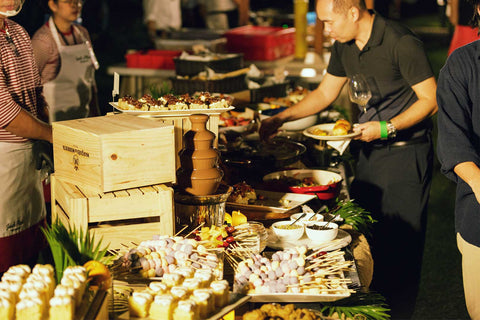 A photo of an outdoor event where guests serving themselves from a buffet table filled with various dishes, desserts, skewers, and a chocolate fountain
