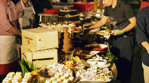 A photo of an outdoor event where guests serving themselves from a buffet table filled with various dishes, desserts, skewers, and a chocolate fountain