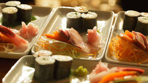 Sushi rolls and sashimi slices neatly plated on 4 white rectangular plates. 
