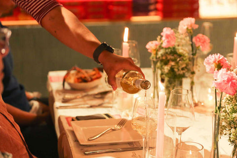 A waiter pouring white wine into a wine glass at a dining table set up with candles, pink flowers, wine glasses, white plates, and cutlery