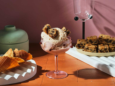 A cup of New York Cheesecake and Cookies Gelato by Chef Miko against a pink background with several cones on the left and square bite-sized cookies on the right