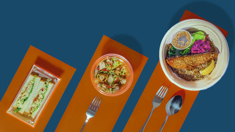 A landscape photo of Joel's place Grab and Go meals on to-go packagings against an orange and navy blue backround with silver spoon and forks on the side