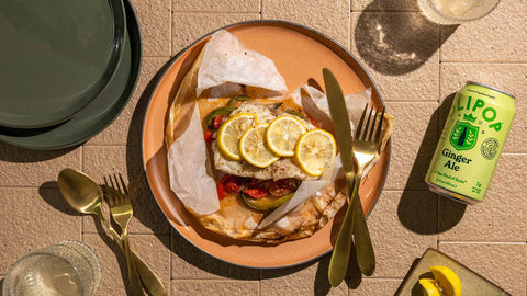 The Deli's Mediterranean Sole en Papillote on top of an orange plate with gold utensils, two green plates, two glasses, and a bottle of Ginger Ale Olipop on the side.
