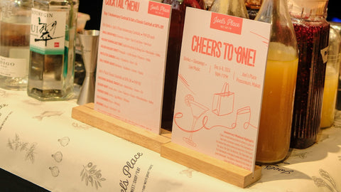 An image of in-store news at Joel's Place, featuring promotional materials for the "Cheers to One!" event celebrating their first anniversary at Proscenium, Makati.