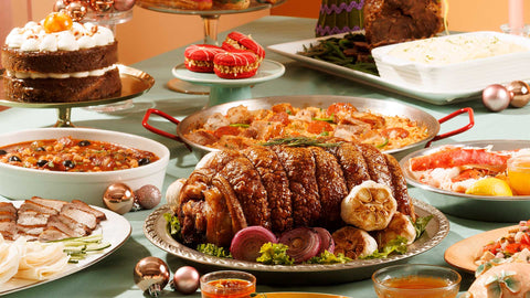 A light green table filled with Christmas holiday meals from Joel's Place including Crispy Crunchy Pork Belly, Festive Callos A La Casa, Party-Ready Meat-Lovers Paella, and Sticky Toffee Cake. 