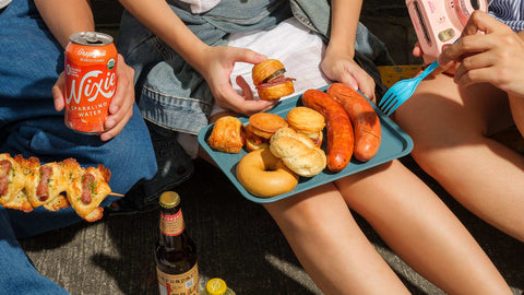A group of people sitting on the ground, enjoying an outdoor street stroll or picnic during summer hangouts, focusing on Joel’s Place Special Orders with croissandwiches, sausages, and refreshing drinks shared among them.