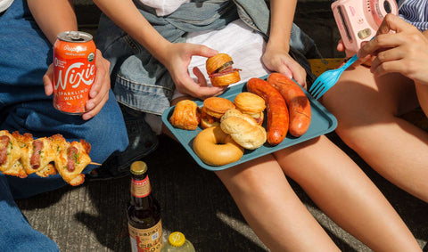 A group of people sitting on the ground, enjoying an outdoor street stroll or picnic during summer hangouts, focusing on Joel’s Place Special Orders with croissandwiches, sausages, and refreshing drinks shared among them.