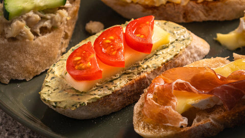 Close-up image of a sliced baguette topped with pesto spread, a slice of mozzarella cheese, and three small slices of cherry tomatoes. It is placed on a green plate with other baguette slices topped with brie cheese and prosciutto.  