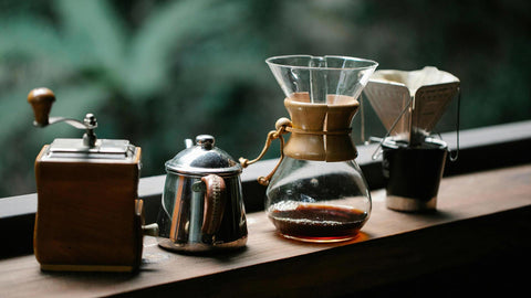 A photo of neatly arranged set of pour-over coffee tools lined up on a countertop, with a n outdoor background filled with greenery and plants