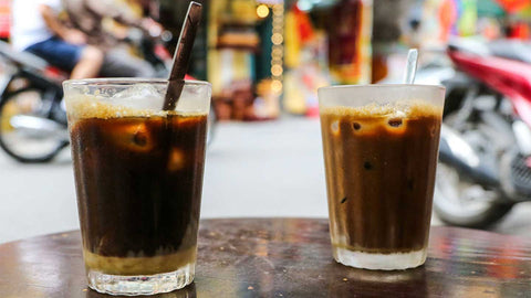 A close-up image of two glasses of Vietnamese iced coffee on a wooden table in a street-side setting in Vietnam.