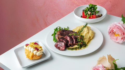A photo of a meal with steak, mashed potatoes, watermelon salad, and a cream-filled pastry, set on a white table with pink flowers