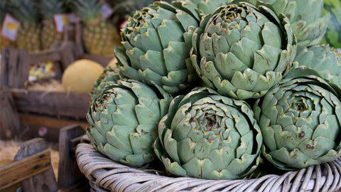 A photo of a basket filled with fresh green artichokes at a market, with pineapples and melons in the background