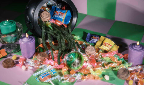 A tipped, overflowing cauldron of candies, chocolates, gummies, and lollipops surrounded by colored candles on a checkered green and purple background