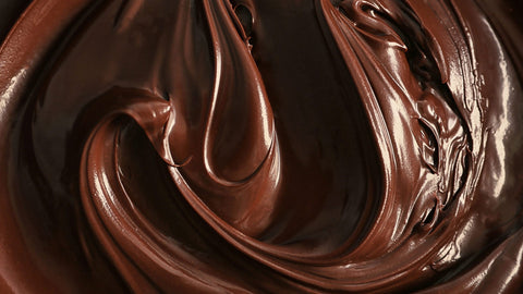 A close-up image of chocolate ganache swirls, showcasing its rich, dark brown color and smooth, glossy texture, featuring a thick, viscous consistency. 