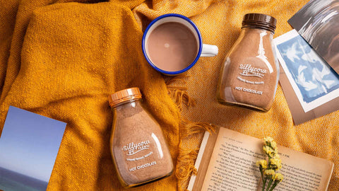 A flat lay photo featuring Sillycow Farms hot chocolate jars, a warm cup of cocoa, an open book, yellow flowers, and a mustard blanket