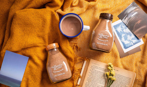 A flat lay photo featuring Sillycow Farms hot chocolate jars, a warm cup of cocoa, an open book, yellow flowers, and a mustard blanket