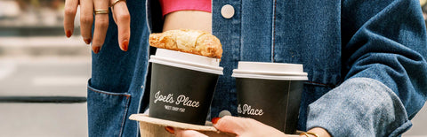 A girl outdoors wearing a denim jacket and holding a carton coffee holder containing two Joel's Place coffee cups, with a pastry on top of one cup