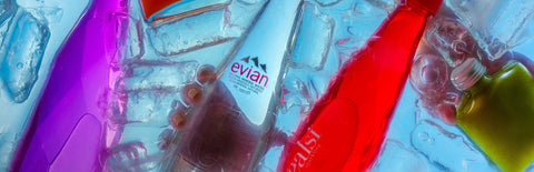 A hand reaching into water filled with ice, Joel's Place powershots, and Healsi water bottles, grabbing a glass Evian water bottle, while another hand grabs a green Joel's Place powershot