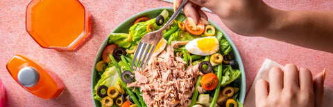 A hand holding a fork over a Joel's Place salad, next to a glass of orange juice and a power shot bottle, on a pink tabletop
