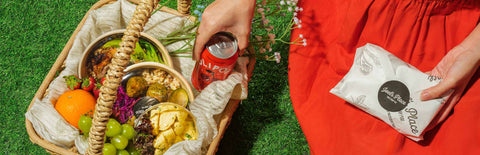 A girl in a red dress taking out a soda can from a picnic basket filled with fruits and Joel's Place packaged meals, while holding a wrapped Joel's Place sandwich in the other hand