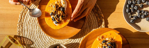 Hands holding a spoon and a Papaya Yogurt Bowl on a wooden plate placed on top of a circular woven placemat next to another Papaya Yogurt Bowl, with a plate of blueberries on the side