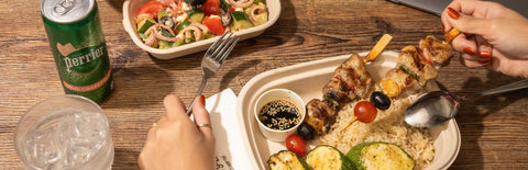Hands holding a fork and meat skewer as she eats a Joel's Place rice meal and salad, with a glass of iced water and a Perrier can