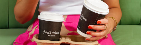 Two Joel's Place coffee cups in a carton coffee holder on the lap of a girl wearing pink polkadot pants and a plain white shirt, sitting on a green couch
