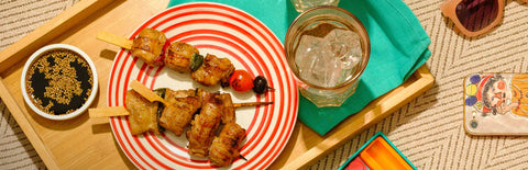 A plate of Joel's Place skewers, a dip with sesame seeds, and a glass of water in a wooden tray next to a mobile phone and peach shades, on top of a patterned tablecloth