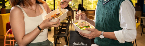 A woman holding a container and a forkful of Shrimp Aglio Olio facing a man holding a container of Salad Nicoise against an office pantry with black Joel's Place takeout bag and two opened canned drinks. 