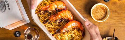 Two hands holding a container filled with Joel's Place Shrimp Aglio Olio. It is against a wooden table with sandwiches, sauces, drinks, and napkins on the side. 